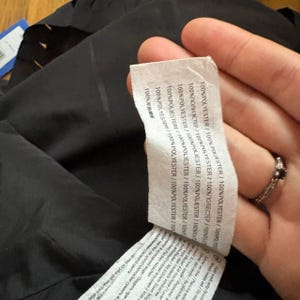 K&ouml;nnte beinhalten: Nahaufnahme eines schwarzen Kleidungsst&uuml;cks mit einem wei&szlig;en Etikett. Das Etikett zeigt Text in mehreren Sprachen, darunter "100% POLYESTER". Das Kleidungsst&uuml;ck scheint aus einem glatten Stoff zu bestehen. Das Etikett wird von einer Hand gehalten.