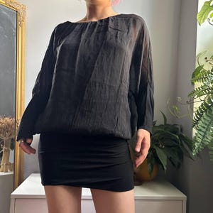 May include: A black long-sleeved blouse with a gathered neckline and a slightly sheer, lightweight fabric, paired with a solid black mini skirt. The blouse features a loose, draped fit with cuffed sleeves, while the skirt has a fitted silhouette.