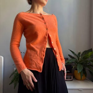 May include: A bright orange long-sleeved cardigan with a wide boat neckline and a front button closure featuring small, light-colored circular buttons. The garment has a soft, textured knit appearance and is paired with a black pleated skirt.