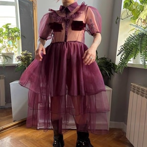 May include: A sheer, deep plum-colored dress featuring short puff sleeves, a collared neckline, and a button-down front. The garment is constructed with multiple layers of ruffled tulle fabric, creating a voluminous, tiered skirt silhouette.