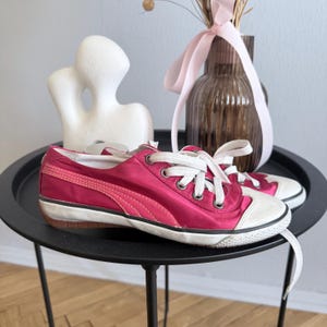 May include: A pair of pink Puma sneakers with white laces and a white toe cap. The shoes are on a black table with a decorative vase and sculpture in the background. The sneakers have a low profile.