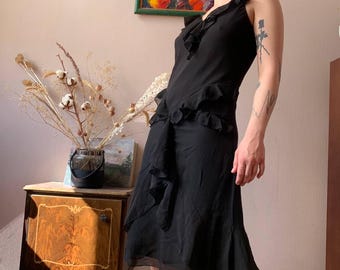 May include: A black dress with a ruffled neckline and asymmetrical hemline. The dress has a flowing, lightweight fabric and a fitted bodice. The dress is sleeveless and has a subtle sheen. The dress is knee-length.