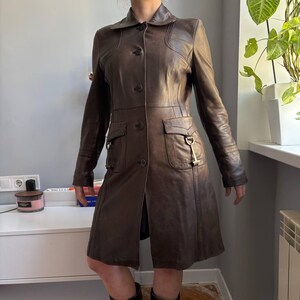 May include: A brown leather coat with a button-up front, two front pockets, and a collar. The coat has a tailored fit and a knee-length hemline. The coat is made of a smooth, supple leather and has a classic, timeless design.