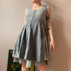Könnte beinhalten: Graues ärmelloses Kleid mit Patchwork-Design. Das Kleid hat ein gestreiftes Oberteil, einen ausgestellten Rock mit gestickten Details und einen geschichteten Saum. Das Kleid hat eine Knopfleiste vorne und einen roten vertikalen Akzent.