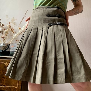 May include: Olive green pleated skirt with three black leather buckle closures. The skirt has a classic design with a flared silhouette. The fabric appears to be a sturdy cotton or blend, suitable for casual wear.