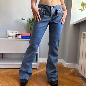 Może przedstawiać: Jasnoniebieskie dżinsy z niskim stanem i rozszerzanymi nogawkami. Dżinsy mają dwie małe przednie kieszenie i zapięcie na guziki. Dżins ma wyblakłe pranie, co nadaje mu wygląd vintage. Dżinsy są noszone z czarnymi butami.