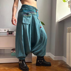 May include: Teal harem pants featuring a high-waisted design with a drawstring closure, a single front pocket, and a multicolored striped waistband detail. The pants have a loose, baggy silhouette that tapers at the ankles, paired with black boots.