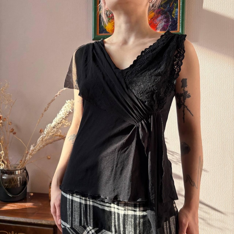 May include: Black sleeveless top with a V-neck and lace detailing. The top has a draped design and a flowing, asymmetrical hemline. The fabric appears to be lightweight and slightly sheer, creating a delicate and elegant look. The top is paired with a black and white plaid skirt.