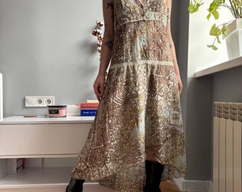 May include: A sleeveless, patterned dress with a V-neckline and spaghetti straps. The dress features a brown and white geometric design, a button-down front, and a flowing, asymmetrical hemline. The model is wearing black boots.