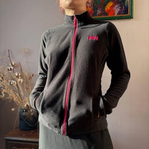 May include: A dark gray fleece jacket with a full-length pink zipper and the pink letters "HH" on the chest. The jacket has a high collar and two front pockets. The model is wearing a dark skirt.