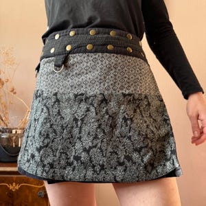 May include: A black and gray patterned skirt with a wide waistband featuring brass-colored button details. The skirt has a geometric pattern at the top and a paisley design at the bottom. A silver-colored ring is attached to the waistband.