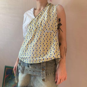 Op de afbeelding: Een wit en geel geprint, mouwloos topje met een wit en blauw geprint stof aan de linkerkant. Het topje wordt gedragen over een lichtblauwe denim short met een riem.