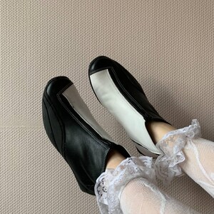 Puede incluir: Un par de botines de cuero negro y blanco con un dise&ntilde;o de color contrastante. Las botas las lleva una persona con calcetines de encaje blanco.