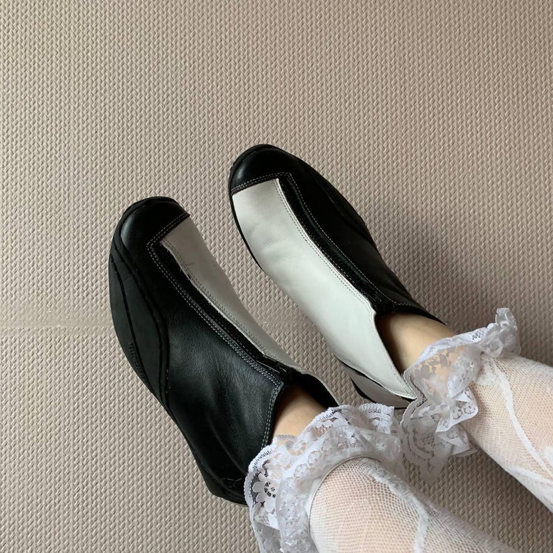 Puede incluir: Un par de botines de cuero negro y blanco con un dise&ntilde;o de color contrastante. Las botas las lleva una persona con calcetines de encaje blanco.