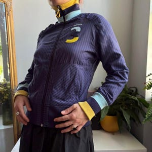 以下が含まれることがあります： ダークブルーのジップアップトラックジャケット。斜めのストライプ模様が施され、イエローとライトブルーのリブ編みトリムが付いたハイカラーが特徴。胸元にはイエローとライトブルーの数字の3のワッペンがあり、その上にAdidasのロゴが配置されている。
