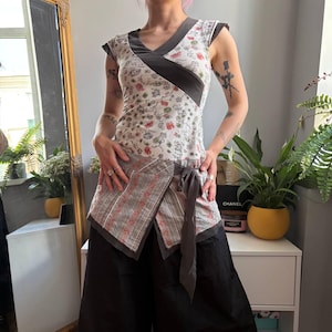 May include: A fitted top featuring a white base with a small floral print, a dark gray V-neckline, and cap sleeves. The lower section has a wrap-style overlay with a gray and white striped pattern, accented by a side tie closure in dark gray fabric.