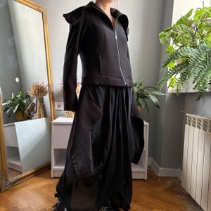 May include: A black long-sleeved jacket with a high collar and a full-length silver zipper, paired with a matching black skirt featuring a draped, asymmetrical hemline and layered fabric panels that create a voluminous, flowing silhouette.