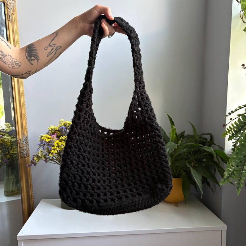 May include: A black crochet shoulder bag featuring an open-knit, textured pattern with a single integrated strap. The bag has a rounded bottom and a wide opening, constructed from thick yarn in a consistent dark shade throughout the entire piece.