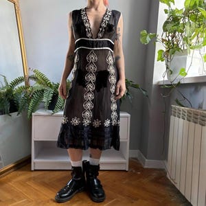 Könnte beinhalten: Ein ärmelloses schwarzes Midikleid mit tiefem V-Ausschnitt, einem breiten horizontalen Taillenband und weißen, vertikal verlaufenden Blumenstickereien auf der Vorderseite. Der Rock hat einen transparenten Saum mit schwarzen Querstreifen.