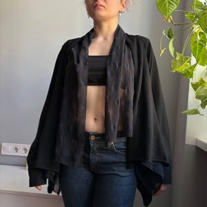 May include: Black open-front cardigan with sheer, patterned panels. The cardigan is paired with a black tube top and dark wash denim jeans. The outfit is styled in a casual, yet fashionable manner.
