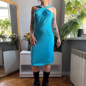 May include: A bright turquoise midi dress featuring a halter neckline with a twisted fabric detail at the chest, a sleeveless design, and a fitted silhouette that falls to the mid-calf, paired with black mid-calf boots.