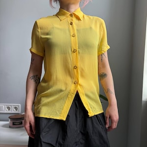 May include: A bright yellow short-sleeved button-down shirt. The shirt has a classic collar and wooden buttons. The fabric appears lightweight and slightly sheer. The shirt is paired with black bottoms.