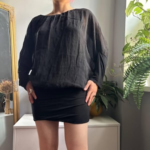 May include: A black long-sleeve blouse with a gathered round neckline and a slightly sheer, textured fabric. The blouse features a loose, draped fit through the torso and sleeves, paired with a solid black, fitted mini skirt with a straight hemline.