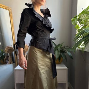 May include: A black wrap-style blouse featuring a high ruffled collar, three-quarter length sleeves with tie cuffs, and a waist tie. The fabric includes subtle embroidered detailing on the front panel, creating a structured and fitted silhouette.