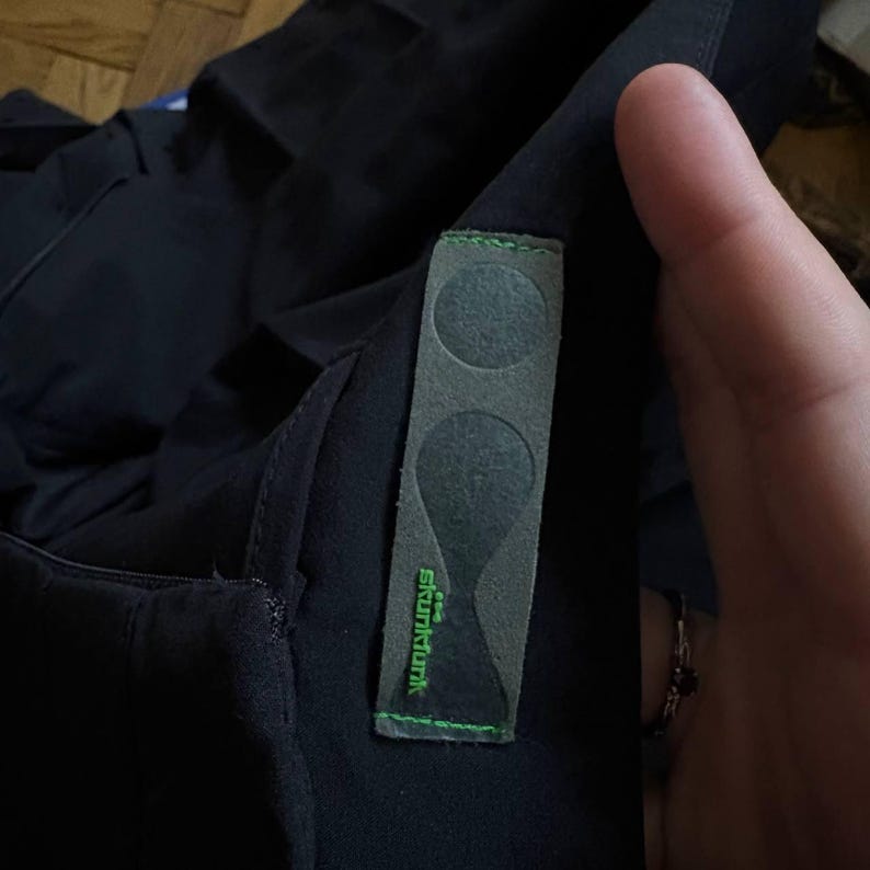 K&ouml;nnte beinhalten: Nahaufnahme eines schwarzen Kleidungsst&uuml;cks mit einem grauen Etikett mit zwei kreisf&ouml;rmigen Formen und dem Text "skunkfunk" in Gr&uuml;n. Das Etikett ist mit gr&uuml;nem Faden gen&auml;ht.