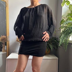 May include: A black long-sleeved blouse featuring a sheer, lightweight fabric with a gathered neckline and a fitted, opaque black mini skirt. The blouse has a loose, draped silhouette with cuffed sleeves, while the skirt sits snugly at the waist.