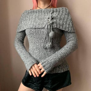 May include: Grey off-the-shoulder sweater with a lace-up detail and pom-poms.