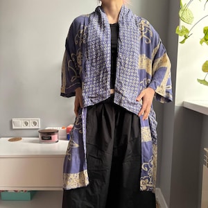 May include: A blue kimono-style robe with a cream-colored pattern. The robe has wide sleeves and a draped front. The pattern includes geometric shapes and floral designs. The robe is worn over a black top and black pants.