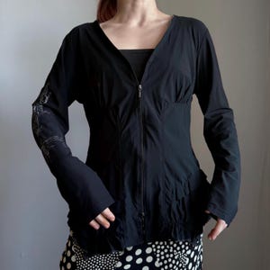Può includere: Giacca nera a maniche lunghe con scollo a V e cerniera a tutta lunghezza. La giacca ha una vita aderente e un fondo svasato. La manica sinistra presenta un design ricamato argentato. La modella indossa un top nero e una gonna a pois bianchi e neri.