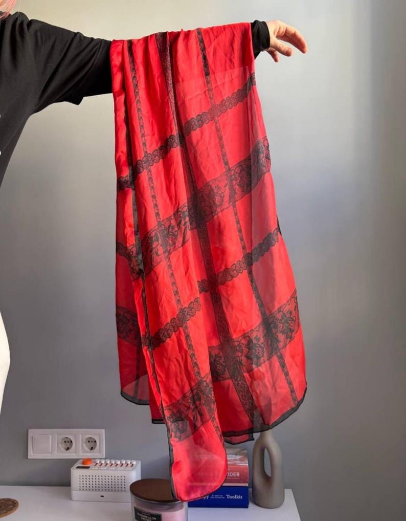 Puede incluir: Una bufanda roja transparente con detalles de encaje negro. La bufanda es rectangular y presenta un patr&oacute;n de rejilla de encaje negro. La tela parece ligera y fluida, adecuada para una variedad de opciones de estilo.