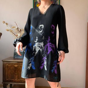 May include: Black dress with long sleeves and a V-neckline trimmed with lace. The dress features a graphic print of a person in blue, white, and purple. The dress has a loose, flowing silhouette.