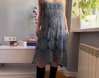 May include: A gray crochet midi dress with spaghetti straps. The dress features a detailed circular pattern throughout the bodice and skirt. The dress is knee-length and has a slight A-line shape. The dress is worn with black boots.