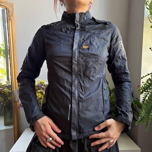 May include: A dark navy blue lightweight jacket featuring a high collar, a central zipper closure with a snap-button placket, and long sleeves with adjustable cuffs. The chest displays a G-Star RAW logo patch and printed text on the sleeve.