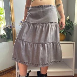 May include: A midi-length skirt in a solid grey color featuring a tiered design with horizontal seams. The waistband and the bottom hem are both detailed with a row of small, dark metal grommets that provide a consistent decorative accent.
