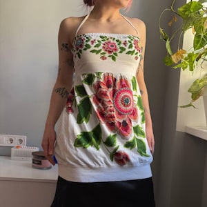 May include: White halter top with a large floral design in shades of pink, red, and green. The top features a fitted bodice and a gathered bottom. The floral pattern is repeated around the neckline. The top is paired with a black skirt.