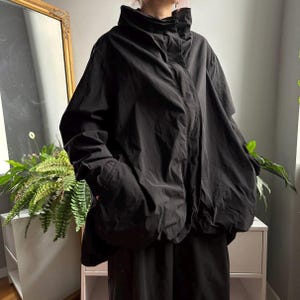 May include: A black oversized jacket with a high collar, long sleeves, and a concealed front button placket. The garment features a voluminous, relaxed silhouette with deep side pockets and a smooth, matte fabric construction.