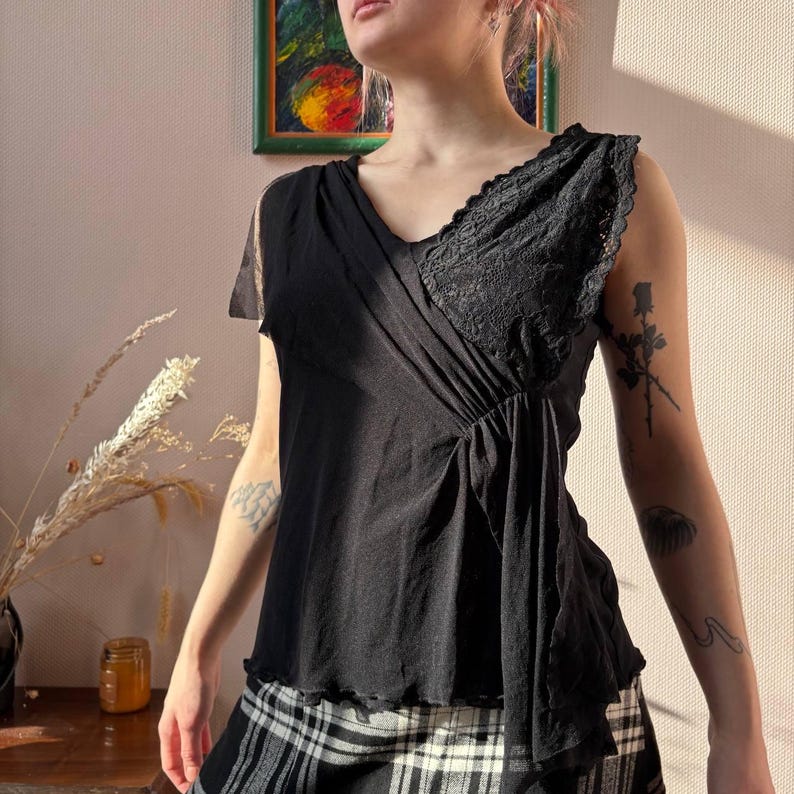 May include: Black sleeveless top with a V-neck and lace detailing. The top has a flowing, asymmetrical design with a gathered waist. The fabric appears to be a lightweight material, suitable for warmer weather. The top is paired with a black and white plaid skirt.