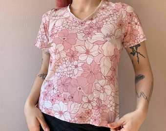 May include: A short-sleeved, V-neck top with a floral pattern in shades of pink and white. The shirt features a delicate, all-over flower design, creating a soft and feminine aesthetic. The neckline and sleeves are trimmed with a lighter shade of pink.