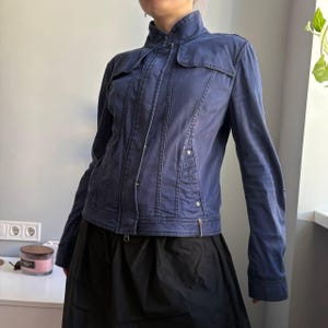 May include: A navy blue jacket with a stand-up collar and button and zipper closure. The jacket has a fitted silhouette with vertical seams and small pockets on the chest. The sleeves are long, and the jacket appears to be made of a durable fabric.