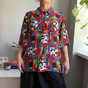 May include: A vibrant, short-sleeved button-down shirt with a bold, geometric pattern. The shirt features a red base with a grid of black, white, and rainbow-colored squares and rectangles. The shirt is paired with a black skirt.