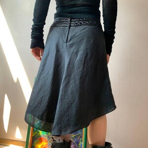 K&ouml;nnte beinhalten: Schwarzer Rock mit asymmetrischem Saum. Der Rock hat ein dekoratives Band am Bund und einen dezenten Glanz. Das Model tr&auml;gt ein schwarzes Oberteil und Stiefel. Der Rock scheint aus leichtem Stoff zu sein.