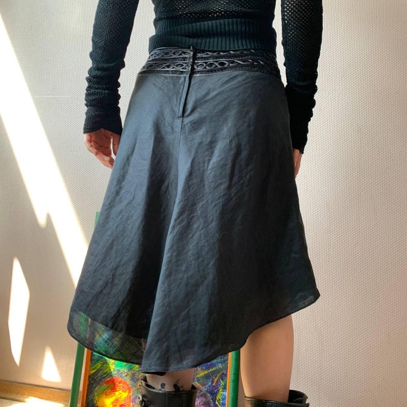 K&ouml;nnte beinhalten: Schwarzer Rock mit asymmetrischem Saum. Der Rock hat ein dekoratives Band am Bund und einen dezenten Glanz. Das Model tr&auml;gt ein schwarzes Oberteil und Stiefel. Der Rock scheint aus leichtem Stoff zu sein.