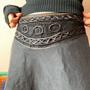 K&ouml;nnte beinhalten: Ein dunkelgrauer Rock mit aufwendig gestaltetem Bund. Der Bund weist eine Reihe dekorativer Elemente auf, darunter Kreise und Webmuster. Der Rock hat eine flie&szlig;ende, ausgestellte Form und der Stoff scheint leicht zu sein.