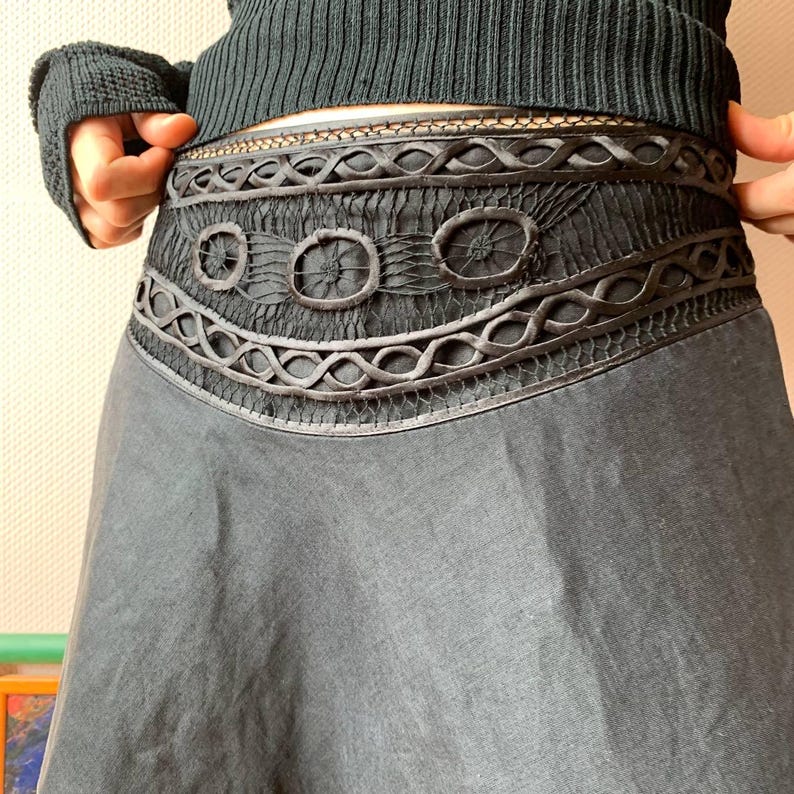 K&ouml;nnte beinhalten: Ein dunkelgrauer Rock mit aufwendig gestaltetem Bund. Der Bund weist eine Reihe dekorativer Elemente auf, darunter Kreise und Webmuster. Der Rock hat eine flie&szlig;ende, ausgestellte Form und der Stoff scheint leicht zu sein.