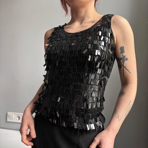 Puede incluir: Top sin mangas negro con lentejuelas. La parte superior sin mangas está cubierta de lentejuelas negras rectangulares, creando un efecto brillante. Tiene un cuello redondo y una silueta ajustada, ideal para una noche.