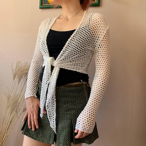 May include: A white, open-weave, long-sleeved cardigan with a tie closure at the waist. The cardigan is worn over a black top and paired with a green skirt. The mesh-like design of the cardigan adds a delicate texture to the garment.
